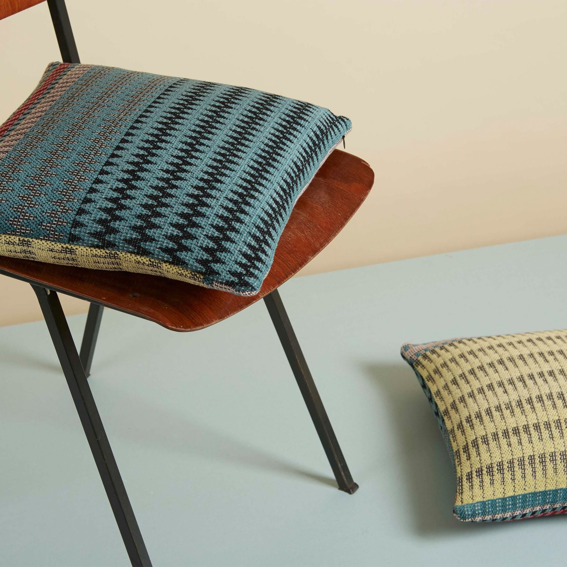Two decorative throw pillows with geometric teal, black, and yellow woven patterns.  One rests on a mid-century modern chair.
