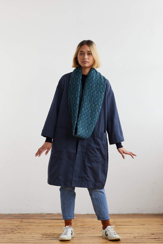 Teal infinity scarf with a subtle repeating pattern, worn by a model over a dark navy coat.