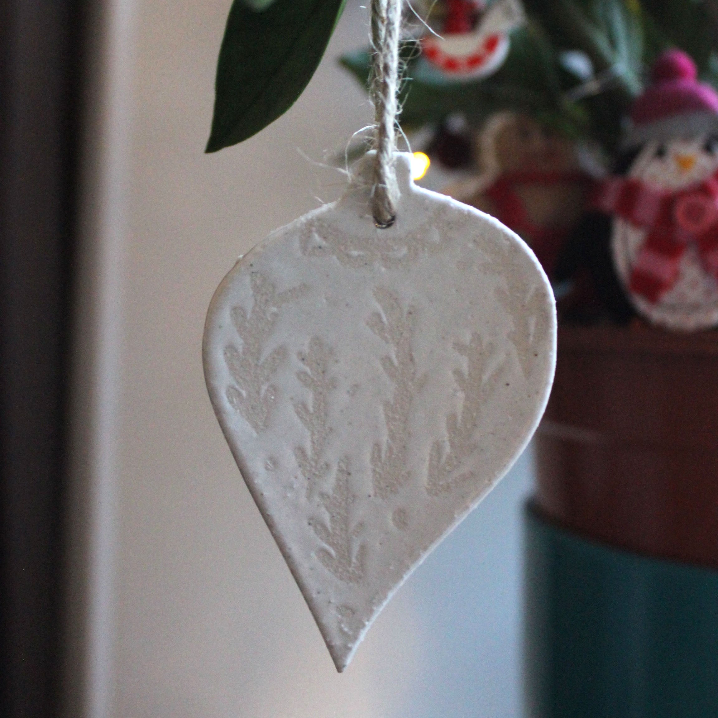 White ceramic Christmas ornament shaped like a leaf, featuring an embossed leaf pattern.  It hangs from a simple twine cord.