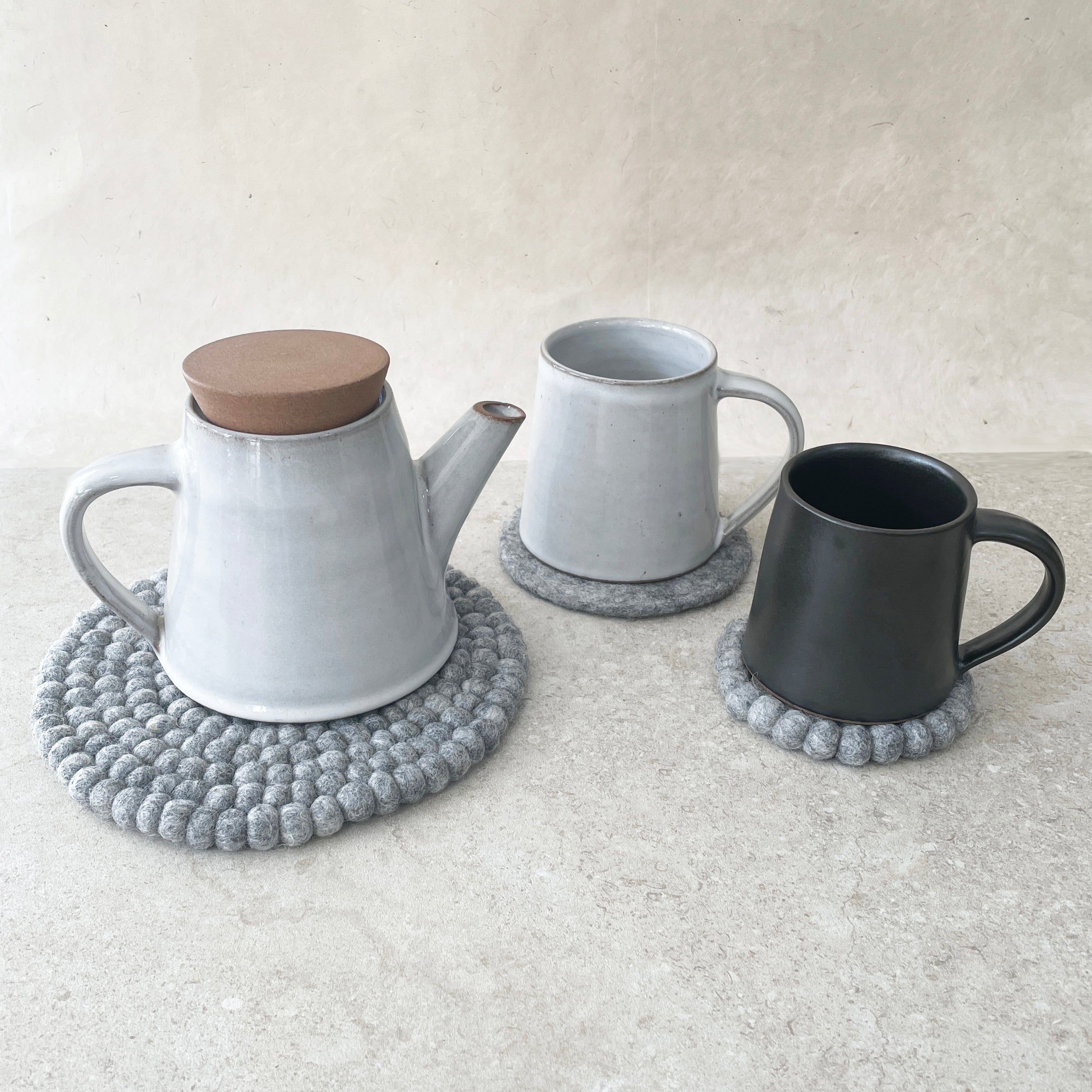 Light gray ceramic teapot with wooden lid and two mugs, one light gray and one black, each resting on a gray felt coaster.