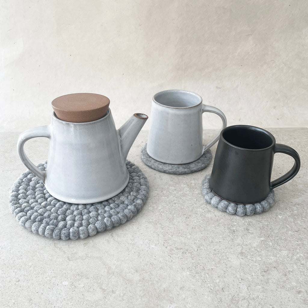 Light gray ceramic teapot with a light brown lid and two mugs, one light gray and one black, each resting on a gray felt