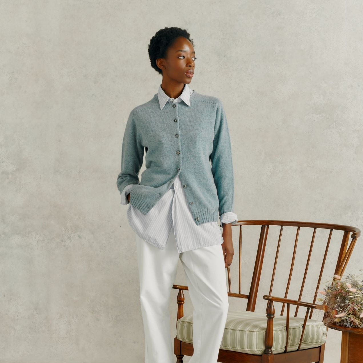 a female model wearing white linen trousers, and a Harley of Scotland Superfine Cardigan - Oribe Green, stood by a wooden chair