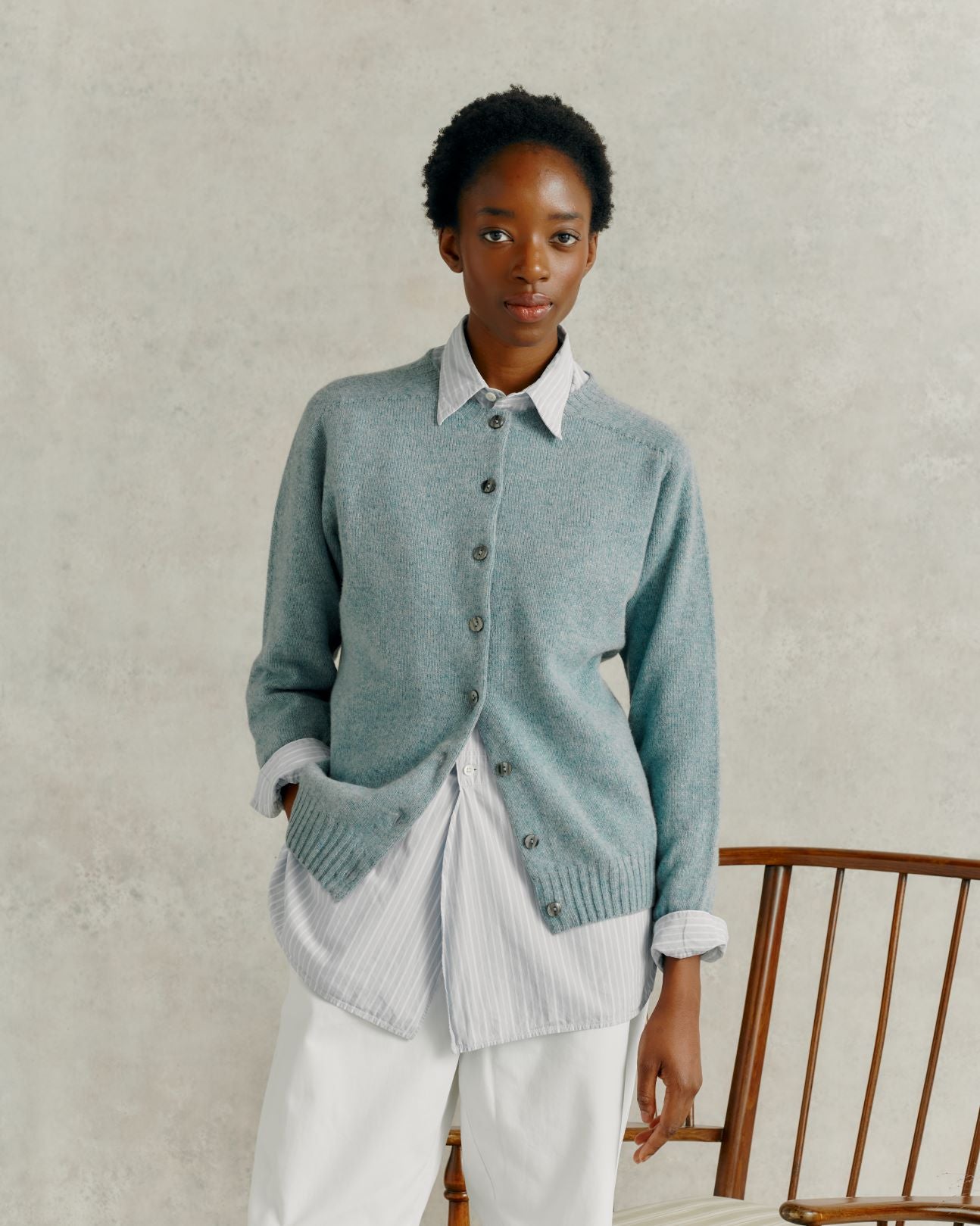 a female model wearing white linen trousers, and a Harley of Scotland Superfine Cardigan - Oribe Green, stood by a wooden chair