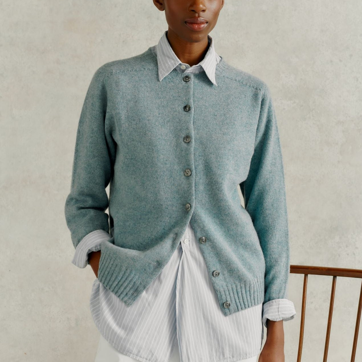 a female model wearing white linen trousers, and a Harley of Scotland Superfine Cardigan - Oribe Green, stood by a wooden chair