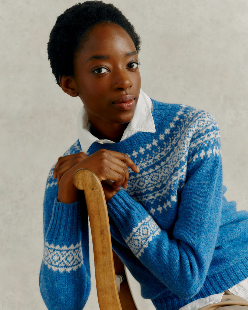 a female model draped over a wooden chair back, wearing a Harley of Scotland Yoke Jumper - Blue Toon & White