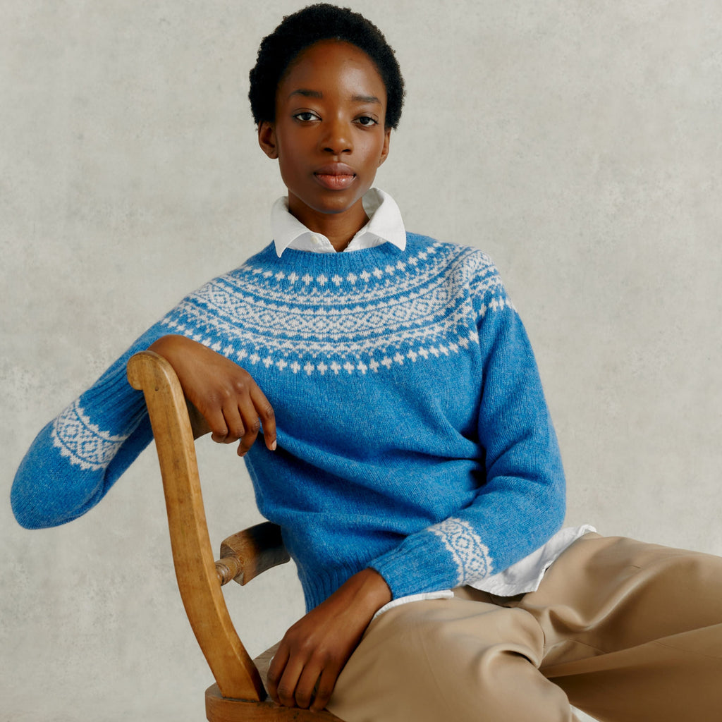 a female model seated on a wooden chair, wearing a Harley of Scotland Yoke Jumper - Blue Toon & White
