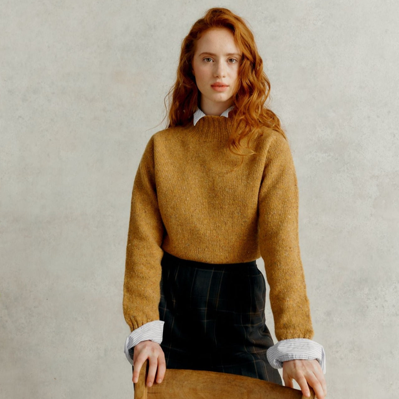 a red haired female model resting forward against a chair, wearing a Harley of Scotland Turtleneck Sweater - Barra