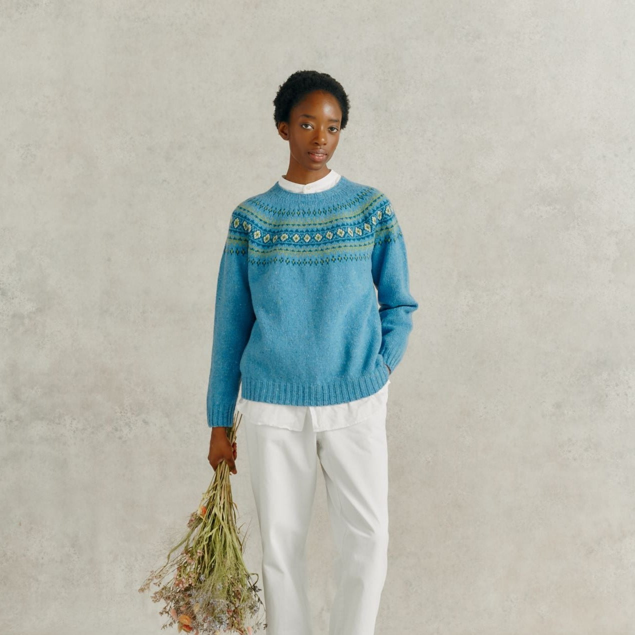 a female model holding a bunch of dried flowers, wearing white linen trousers and a Harley of Scotland Fairisle Jumper - Coll