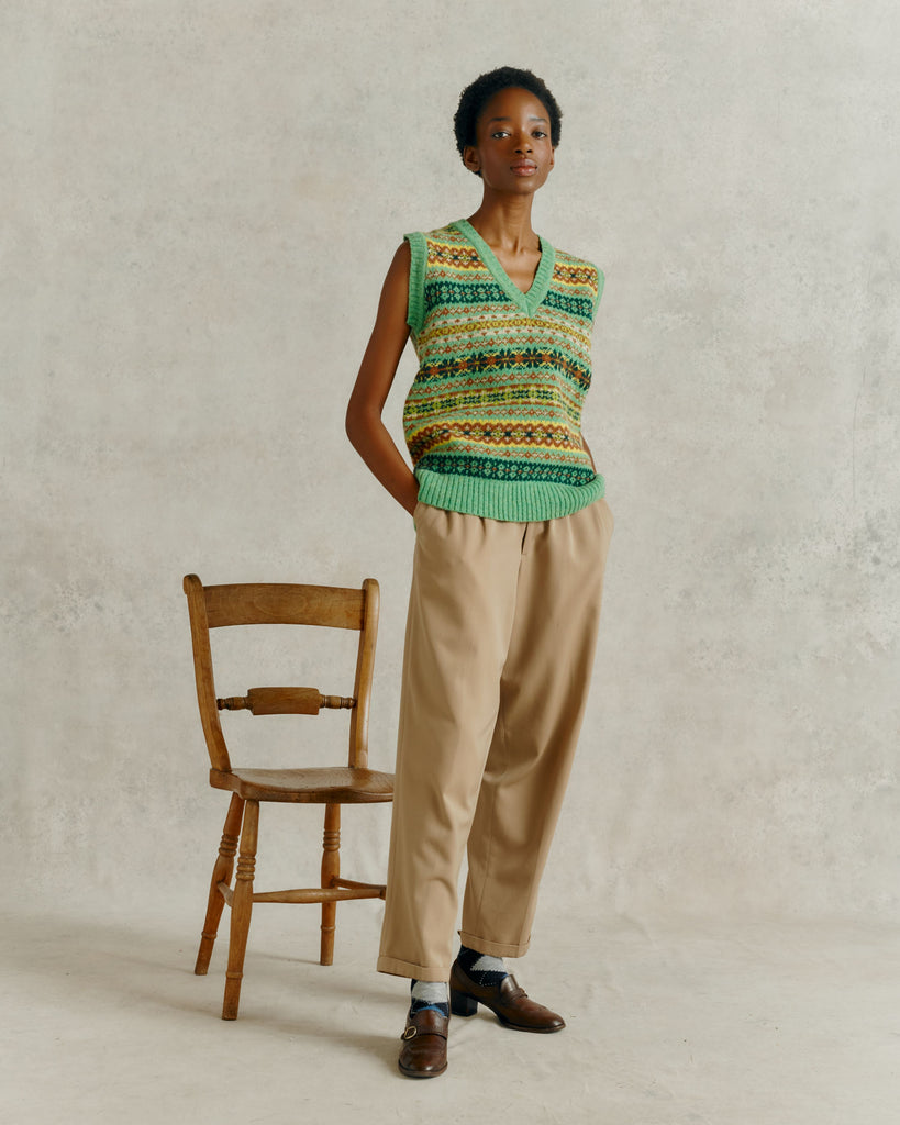 a female model with hands behind back, wearing a Harely of Scotland Fairisle Slipover - Mint LA,  standing in front of a wooden chair