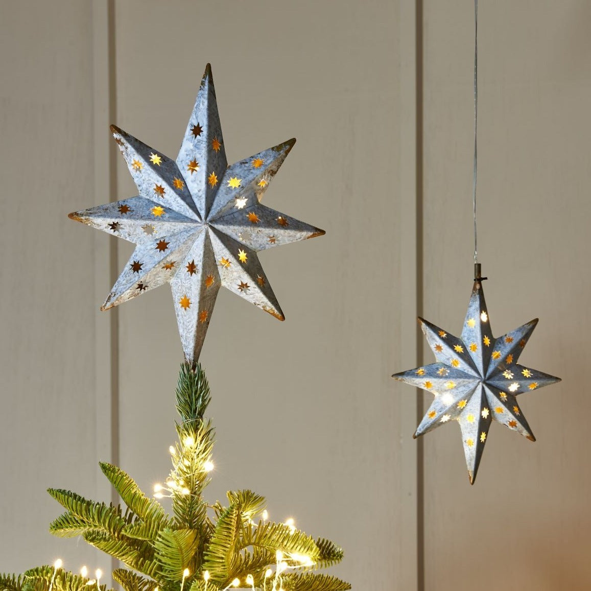 Two illuminated, metallic gray, eight-pointed star tree toppers with gold detailing.  One adorns a Christmas tree.