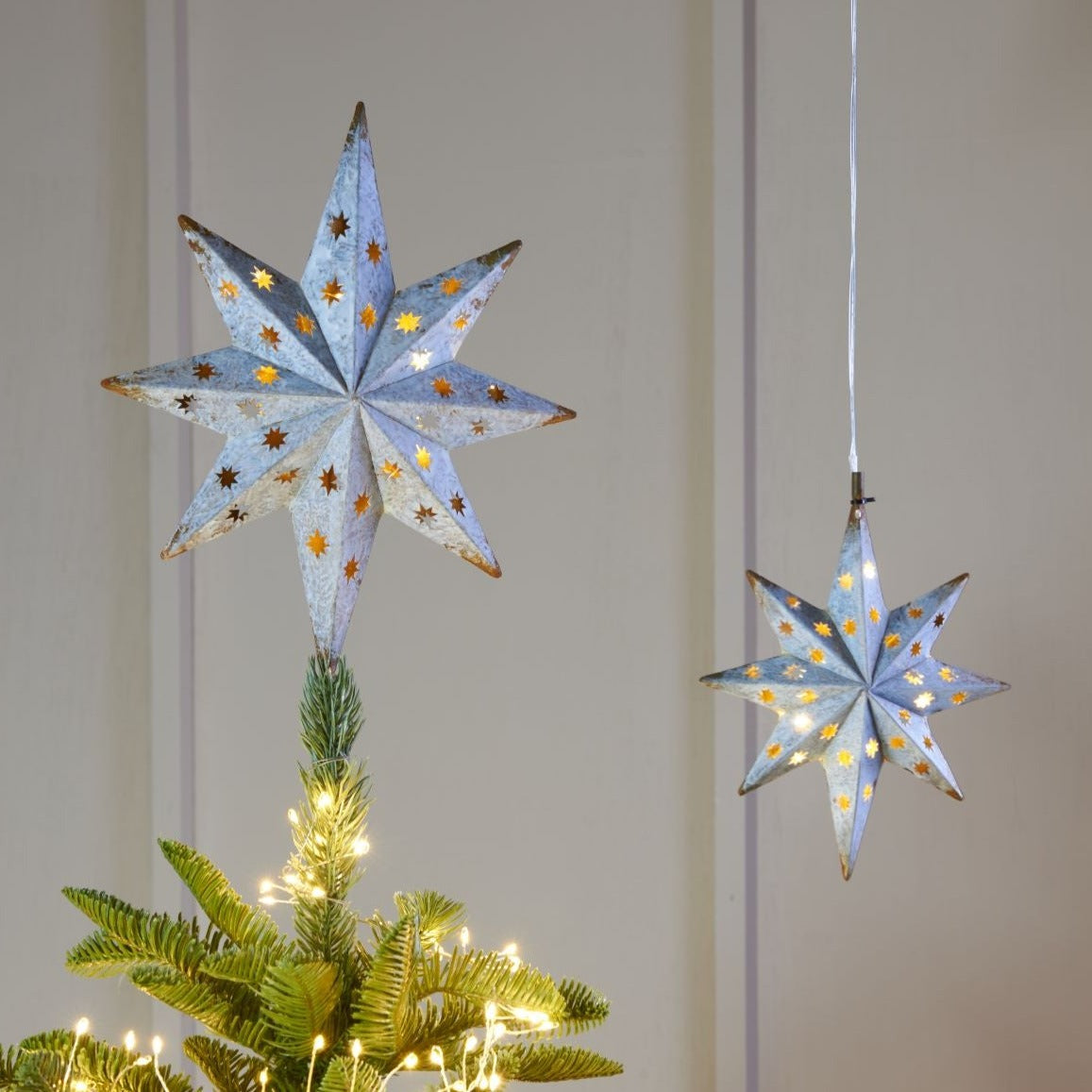 Two illuminated, eight-pointed metal star tree toppers; one atop a Christmas tree, the other hanging.