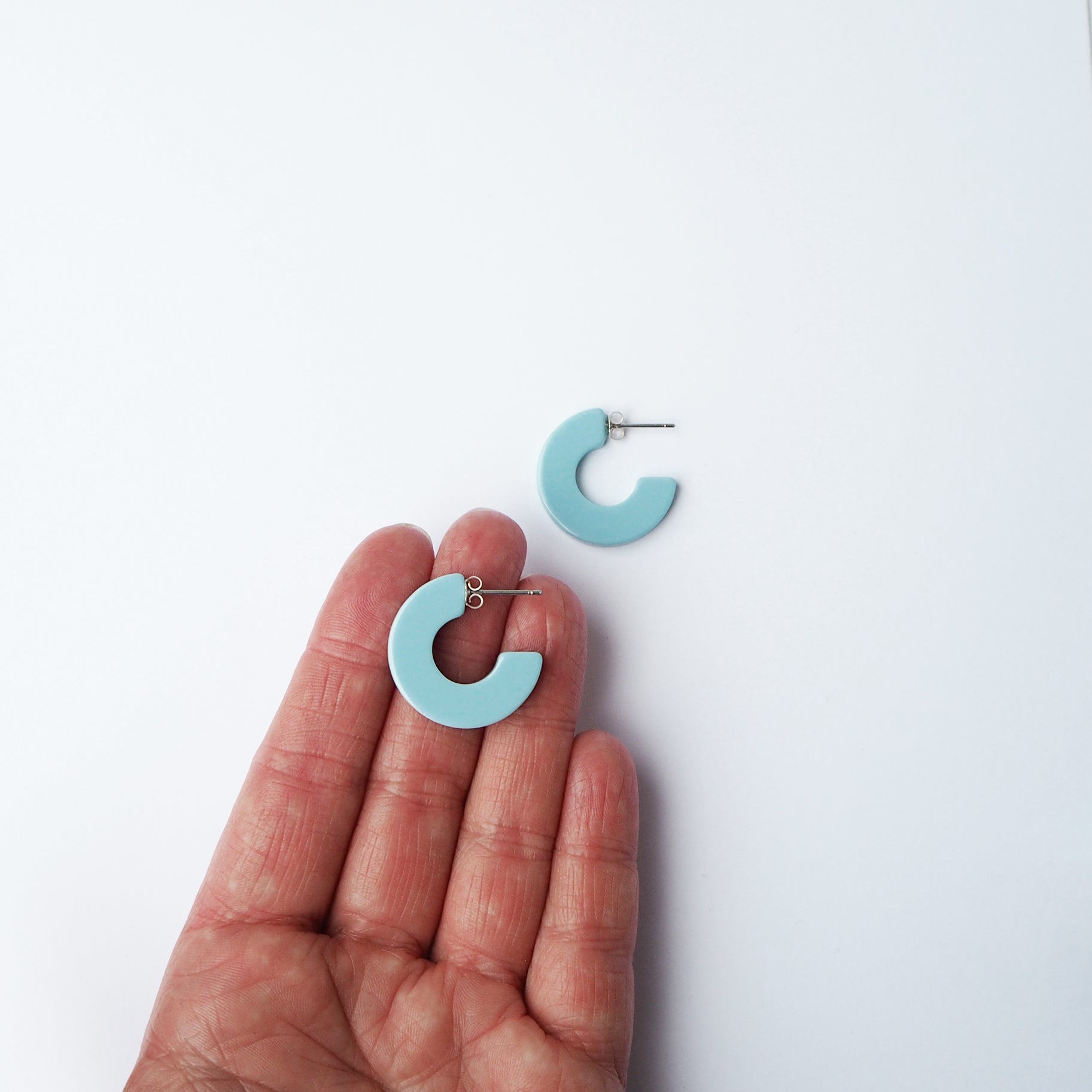 Pair of light blue, semi-circular hoop earrings.  One earring is held in a hand; the other rests on a white surface.