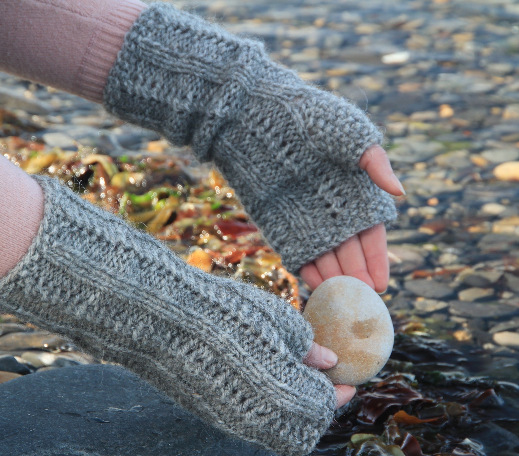 Pair of grey hand-knitted fingerless gloves, textured with cable knit pattern, holding a smooth, light-brown stone.