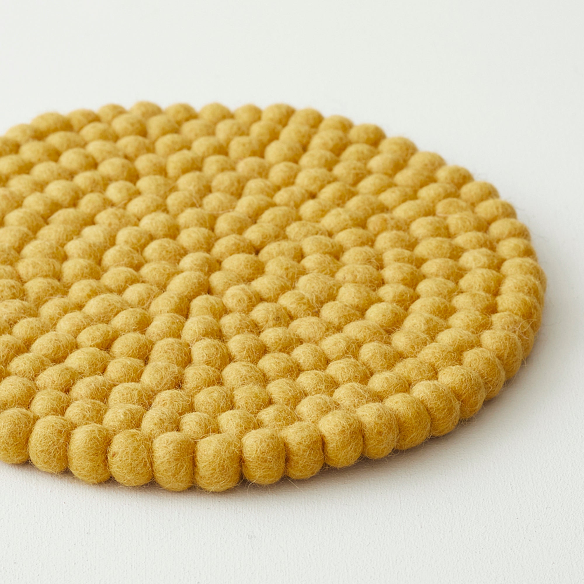 Mustard yellow round felt coaster with a textured surface of small, densely packed pom-poms.