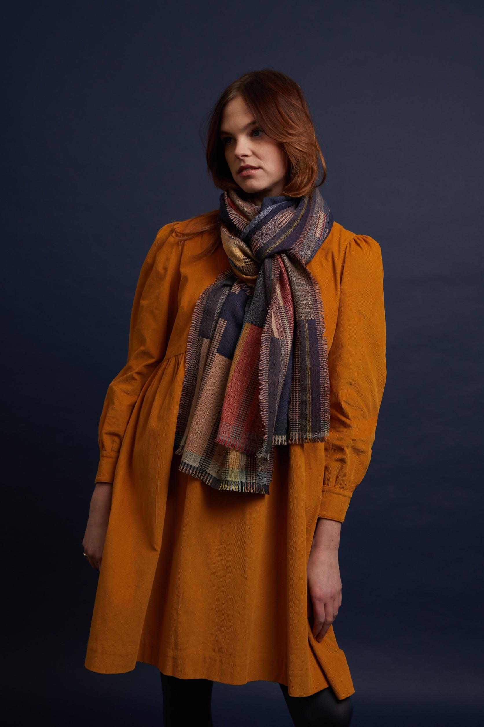 Multicolored woven scarf with a frayed edge, featuring a blend of navy, rust, beige, and mustard yellow in a plaid pattern.
