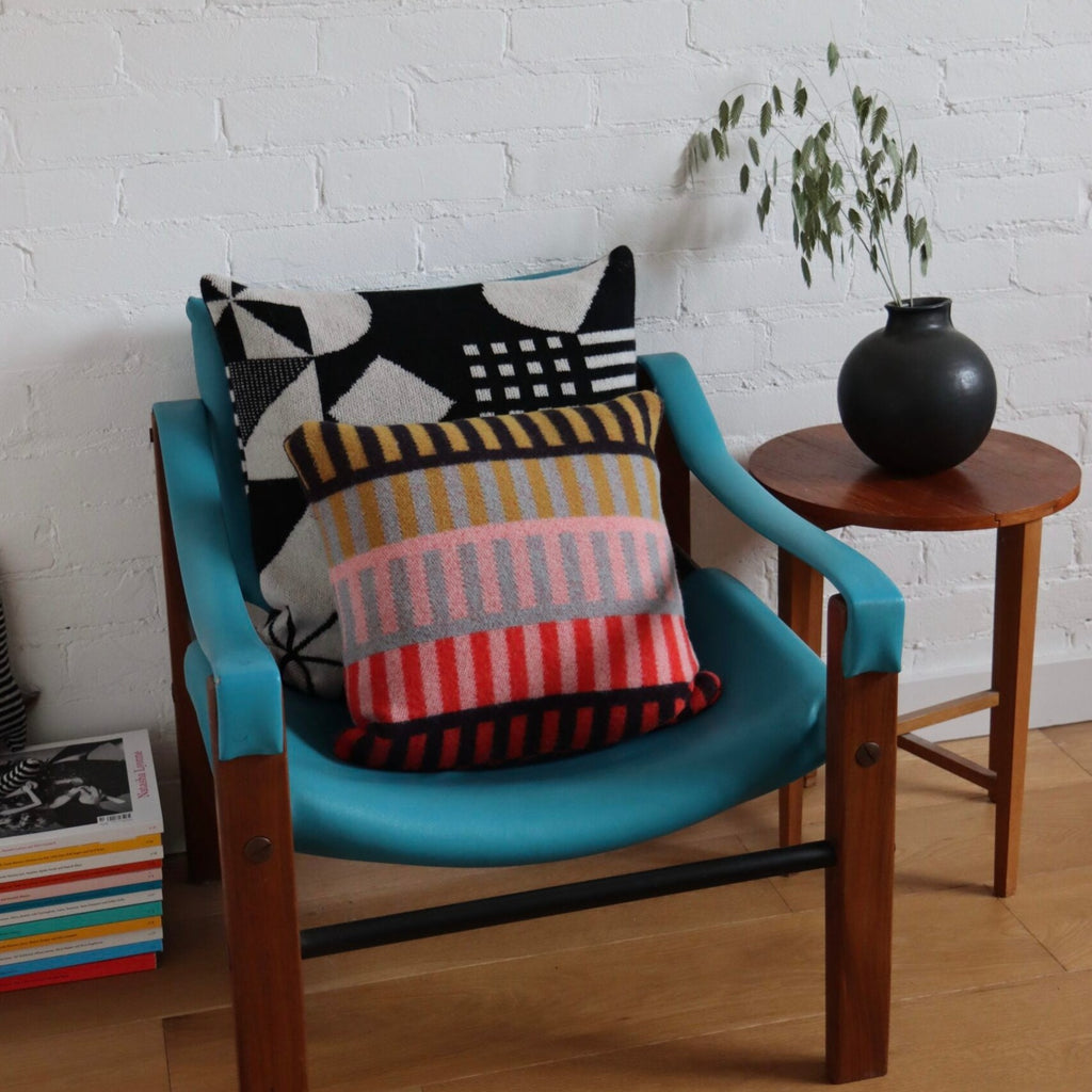 A turquoise chair features two cushions by Donna Wilson: one black and white, the other with stripes in pink, orange, and gray.