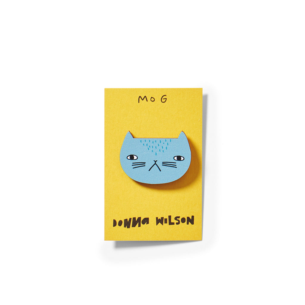 A blue cat brooch on a yellow card, labeled "MOG," by Donna Wilson. Simple and playful design.