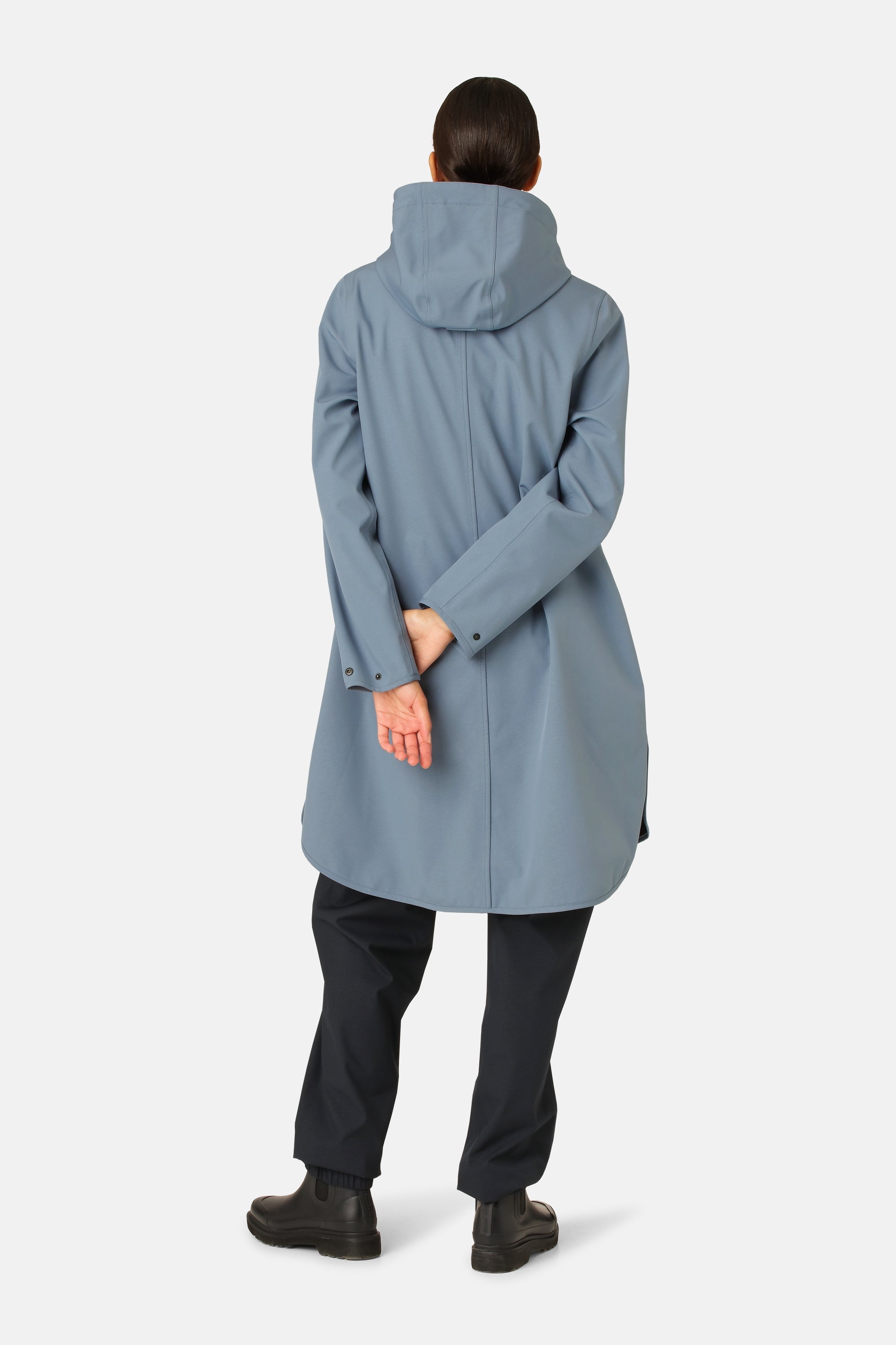 Rear view of a woman wearing a light blue Rains long hooded raincoat, dark pants, and black ankle boots against a white