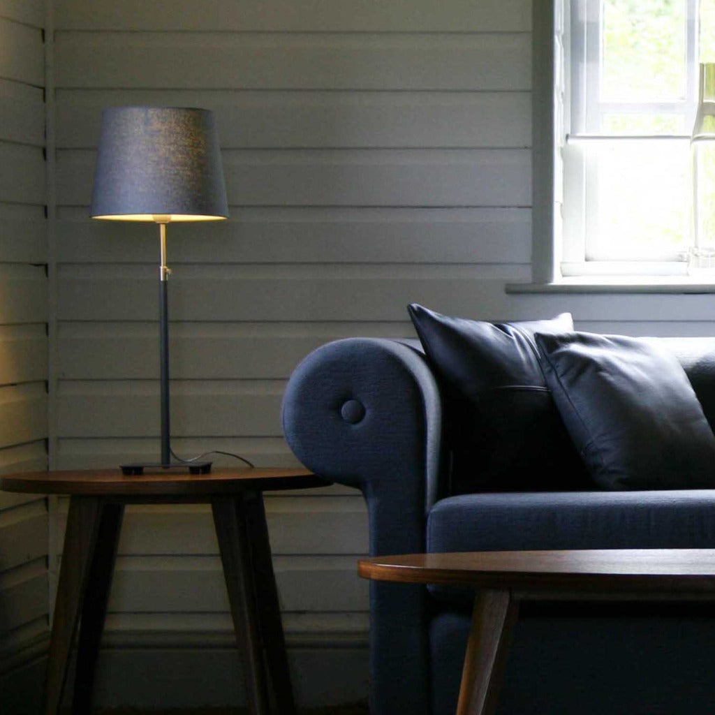 Dark gray table lamp with a cylindrical shade on a small wooden side table, next to a dark blue sofa.