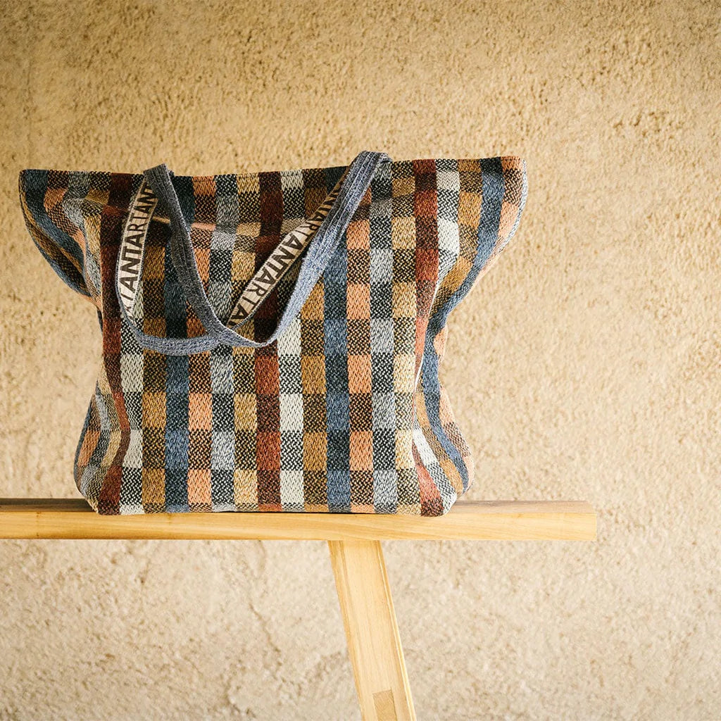 Large woven tote bag with a gingham pattern in muted blues, browns, and creams.  The bag has gray fabric handles with a logo.