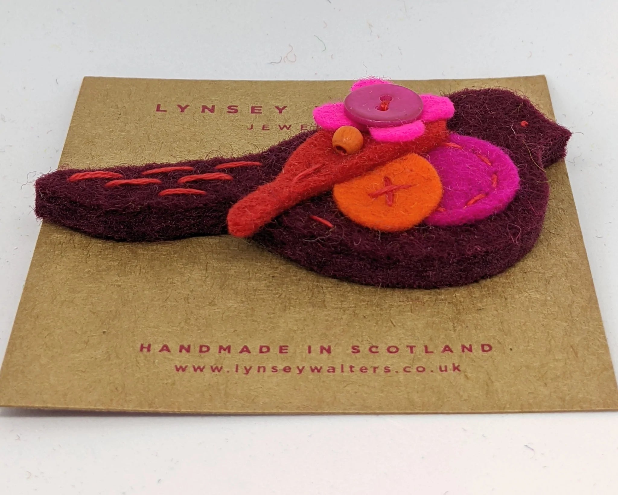Handmade felt bird brooch in maroon, pink, and orange, featuring a button and bead accents.