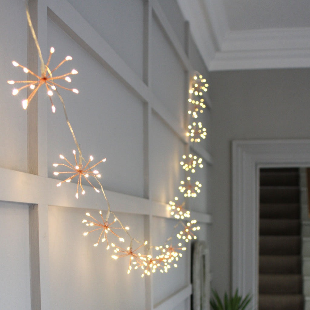 Rose gold firework string lights with warm white LEDs.  The lights are arranged in a curved line along a wall.