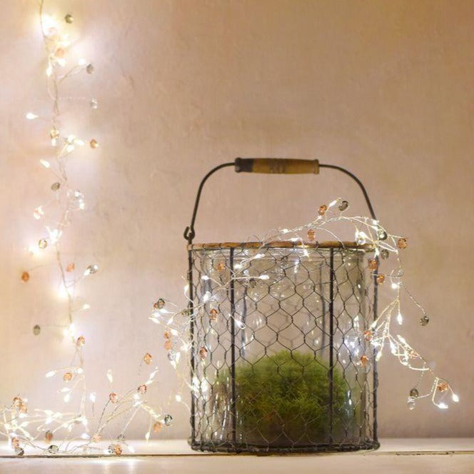 Silver wire string lights with small, warm-toned beads, draped around a wire-mesh container holding moss.