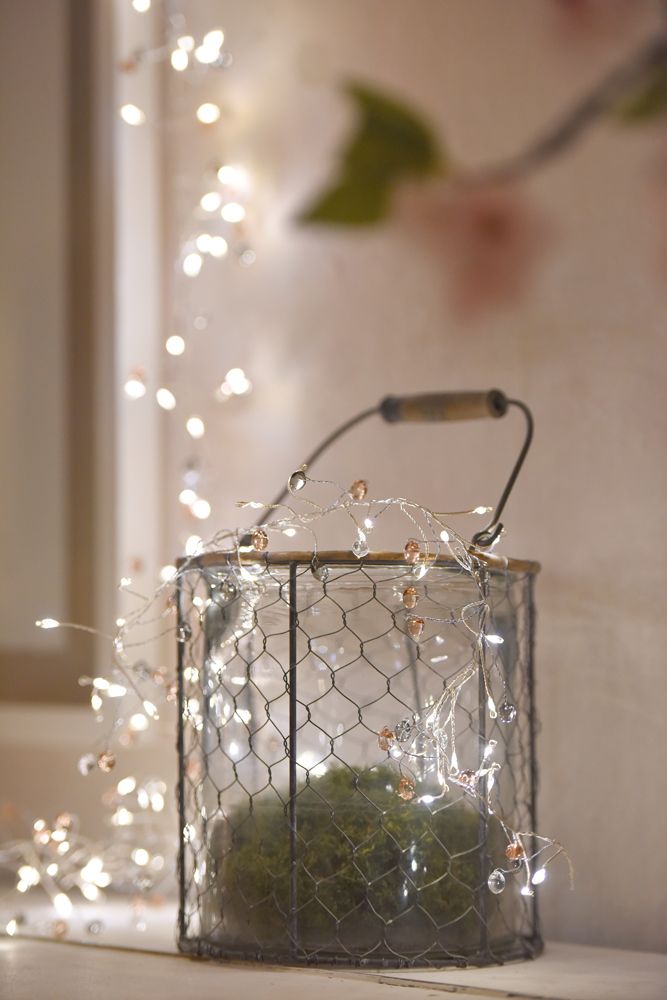 Copper wire string lights with clear and rose-colored beads, arranged in a wire-mesh container filled with moss.