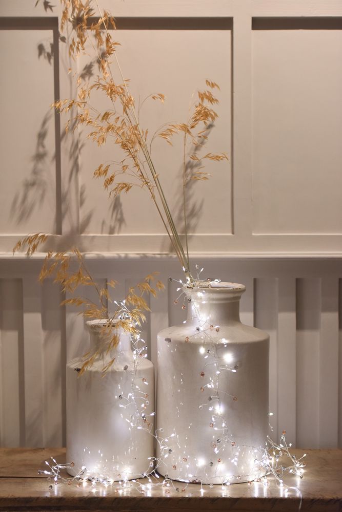 Silver and rose gold beaded fairy lights draped around two white ceramic vases, accented with dried wheat stalks.