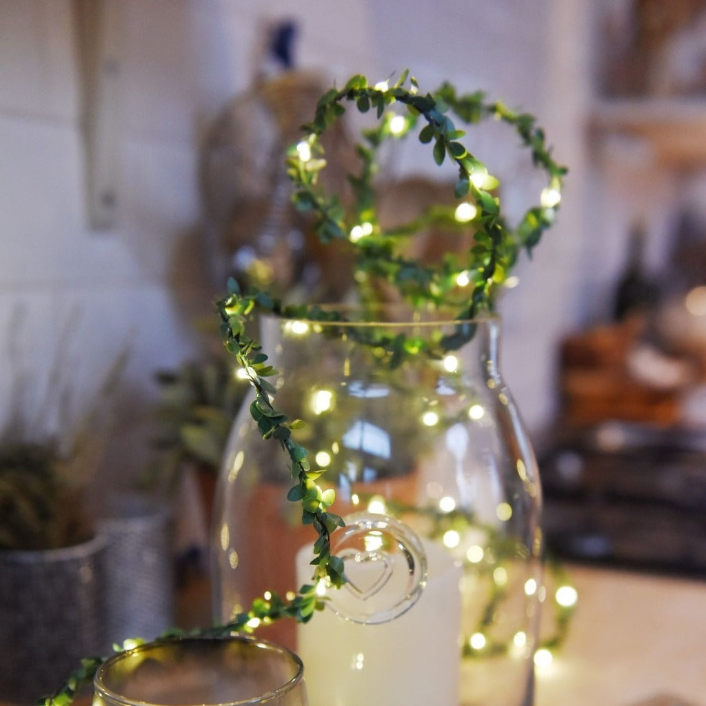 LED string lights wrapped around a clear glass vase;  small green leaves adorn the lights.