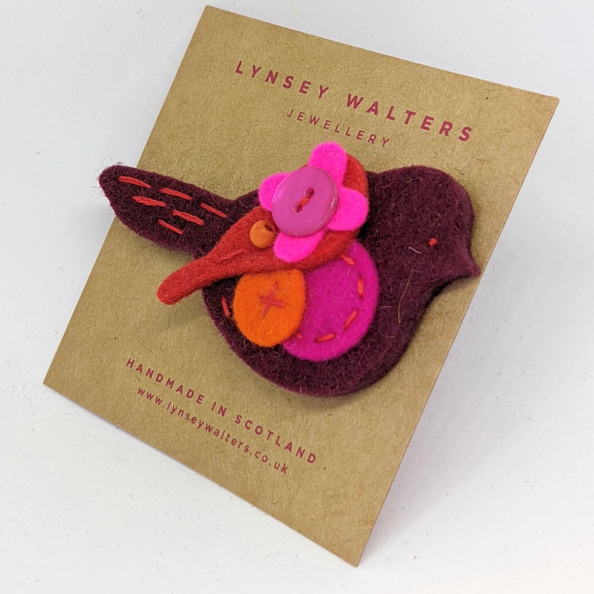 Handmade felt bird brooch in maroon, pink, orange, and red. Features decorative stitching and a pink button flower.