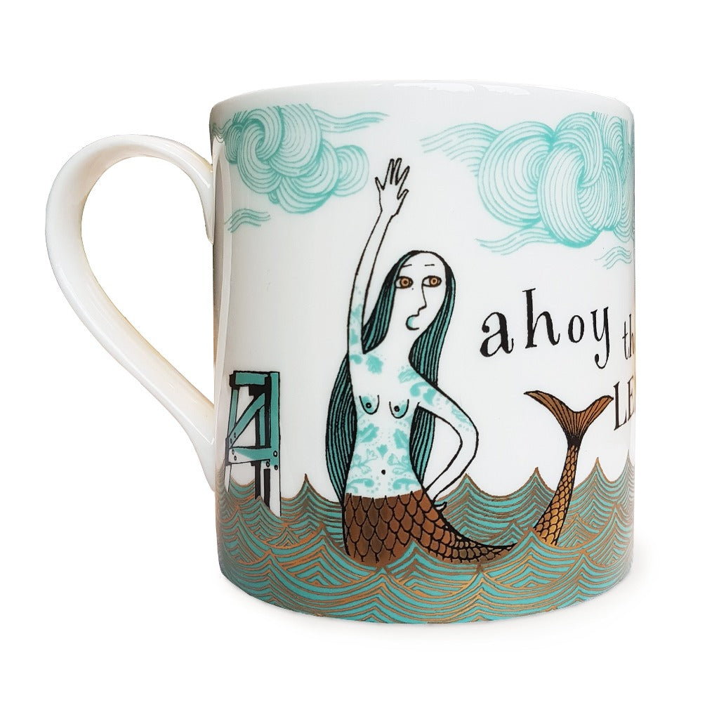White mug featuring a stylized mermaid illustration with teal and gold details; text "ahoy there" is partially visible.