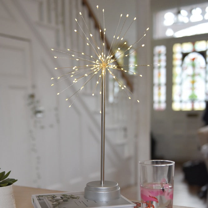 Silver metal table lamp with numerous small, warm-white LED lights radiating outwards, resembling a firework display.