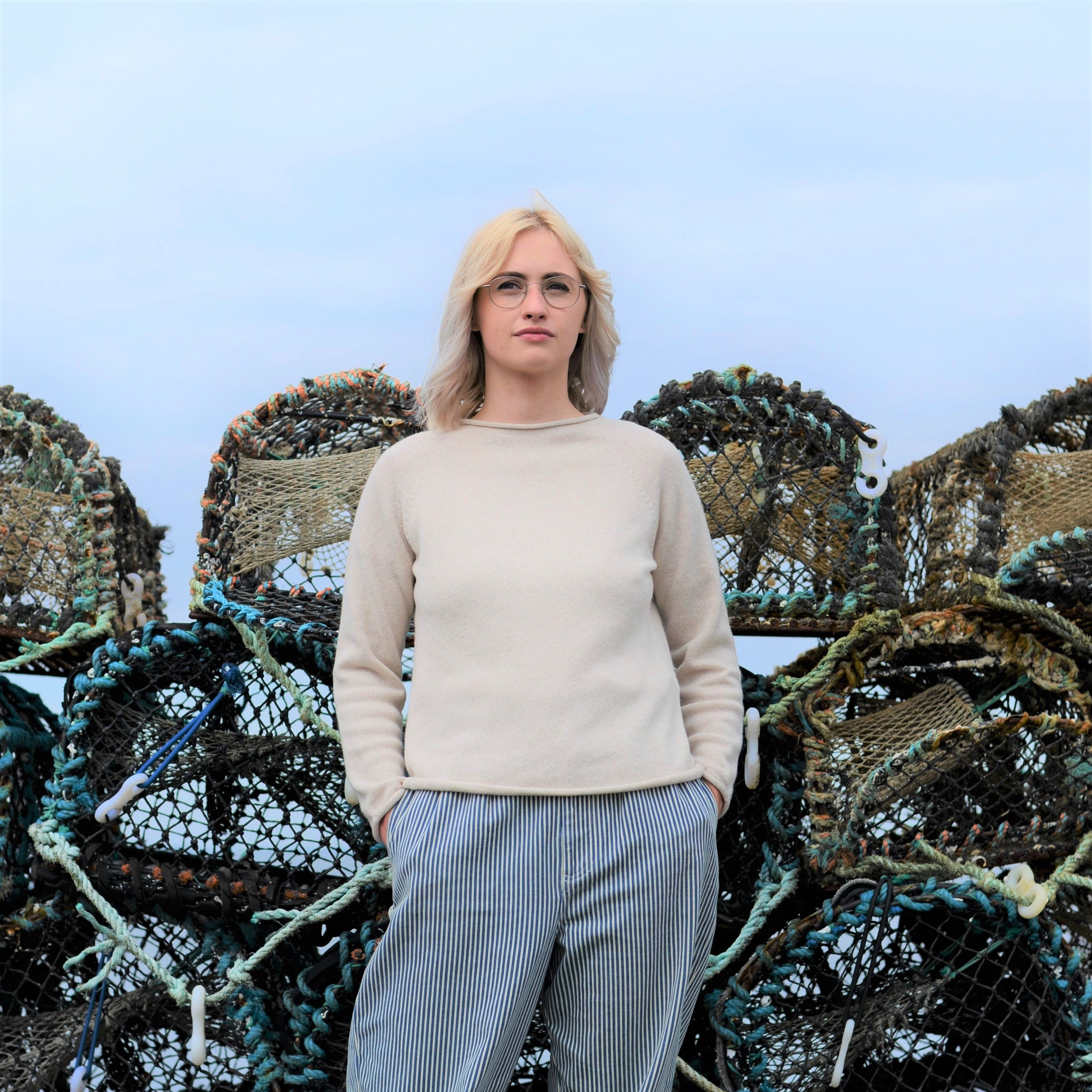 Woman wearing a cream-colored sweater and blue and white striped pants stands in front of a stack of weathered fishing traps.