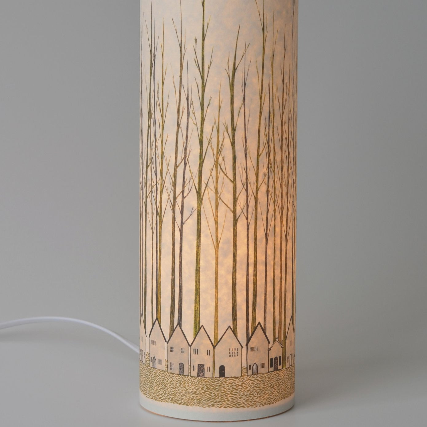 Cylindrical table lamp with a light-colored shade featuring an illustration of bare trees and small houses at the base.